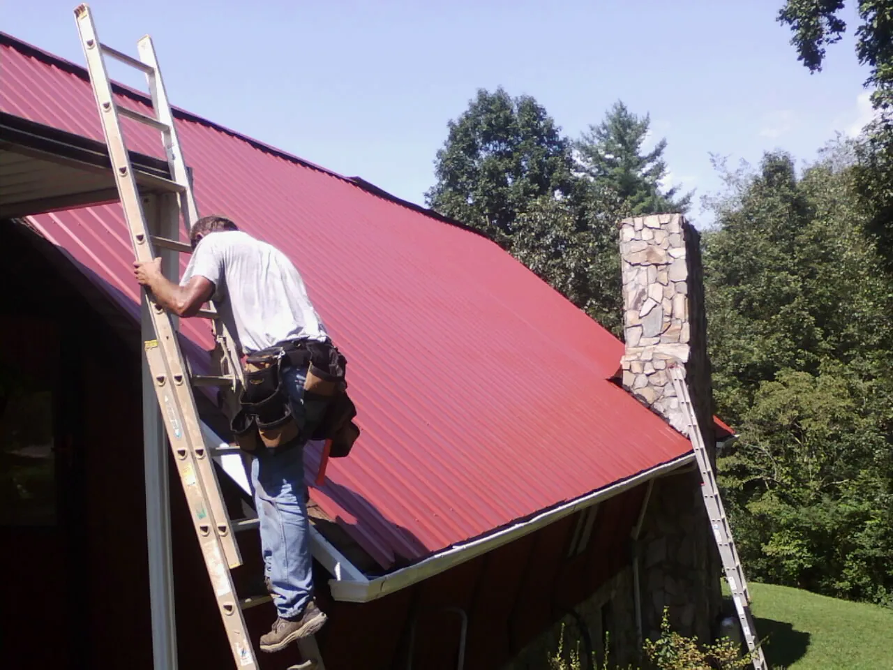 Roofer on a ladder beside a red metal roof and stone chimney — Soffit & Fascia Repair by Roofing Brothers in Random Lake